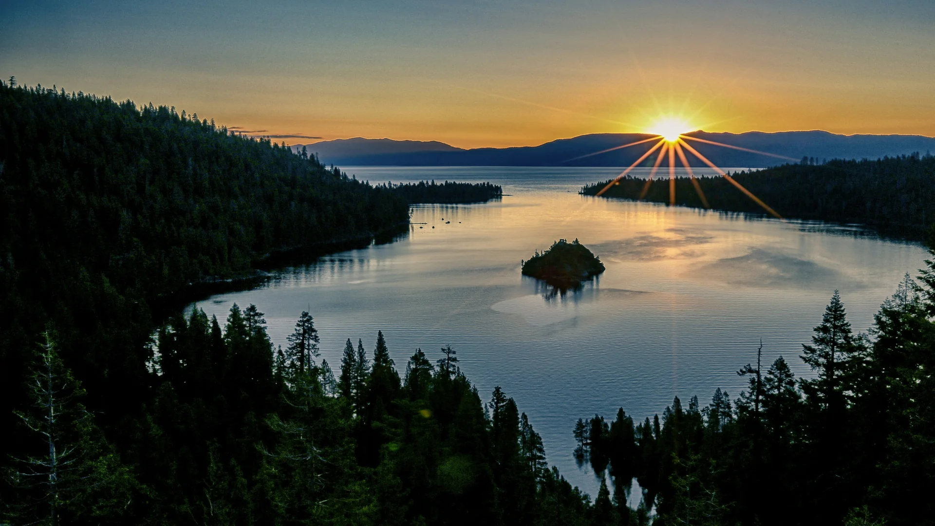 The sun low over the horizon at Emerald Bay on Lake Tahoe, with Fannette Island dark on the glowing water and pine forest framing both sides