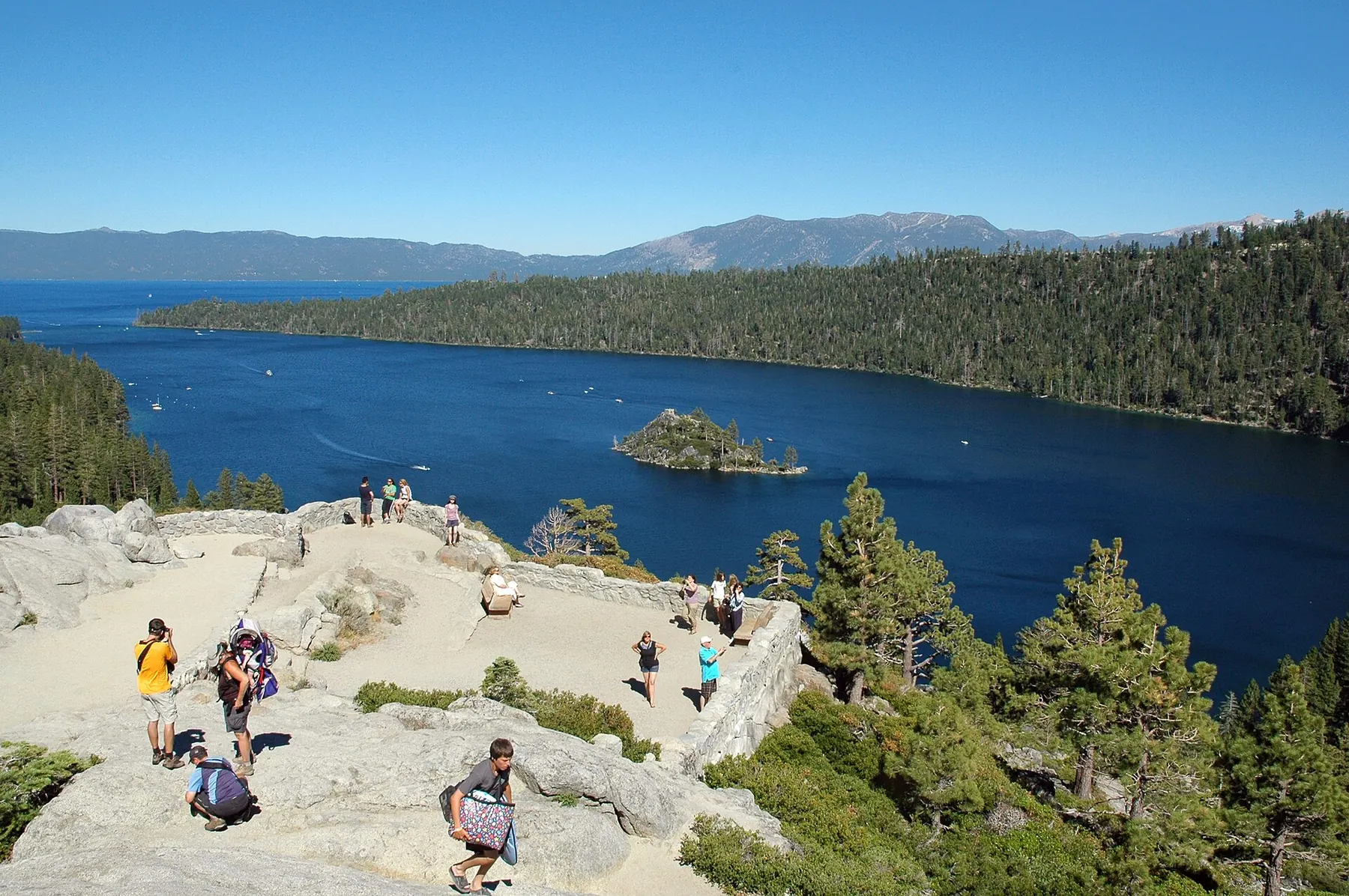 Visitors on a granite terrace at the Emerald Bay lookout above deep blue water, with Fannette Island and forested shores beyond